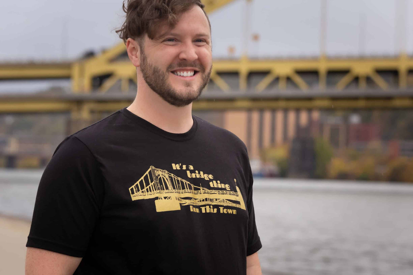 Man standing in front of a river and bridge wearing a black crew neck t-shirt with gold distressed graphic image of a bridge text it's a bridge thing above the bridge, text run this town below the bridge