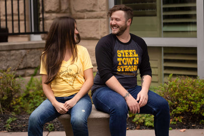 A man and a woman sitting on a bench smiling at each other wearing Burgh Swag shirts 