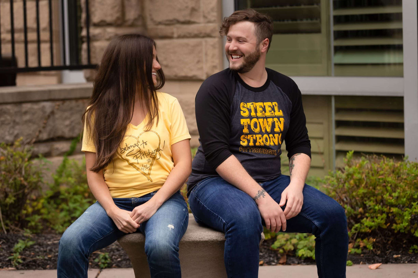 A man and a woman sitting on a bench smiling at each other wearing Burgh Swag shirts 