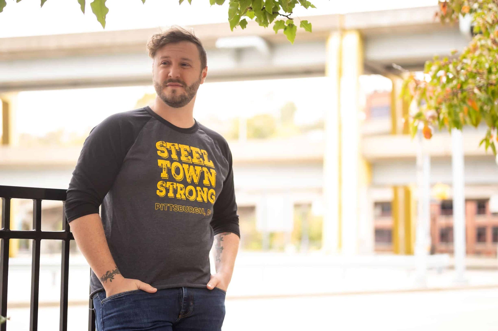 Man leaning against a railing with a blurred city background wearing a baseball tee with gray body and black sleeves with yellow and black distressed graphic Steel Town Strong Pittsburgh PA on front