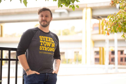 Man leaning against a railing with a blurred city background wearing a baseball tee with gray body and black sleeves with yellow and black distressed graphic Steel Town Strong Pittsburgh PA on front