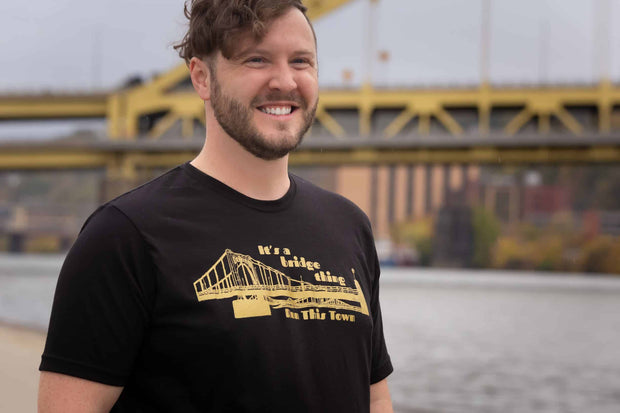 Man standing in front of a river and bridge wearing a black crew neck t-shirt with gold distressed graphic image of a bridge text it's a bridge thing above the bridge, text run this town below the bridge 