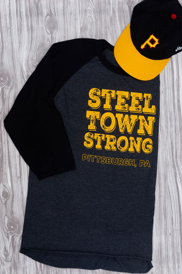 Baseball tee with gray body and black sleeves with yellow and black distressed graphic Steel Town Strong Pittsburgh PA on front folded with a Pittsburgh Pirates baseball cap against a gray wood texture background 